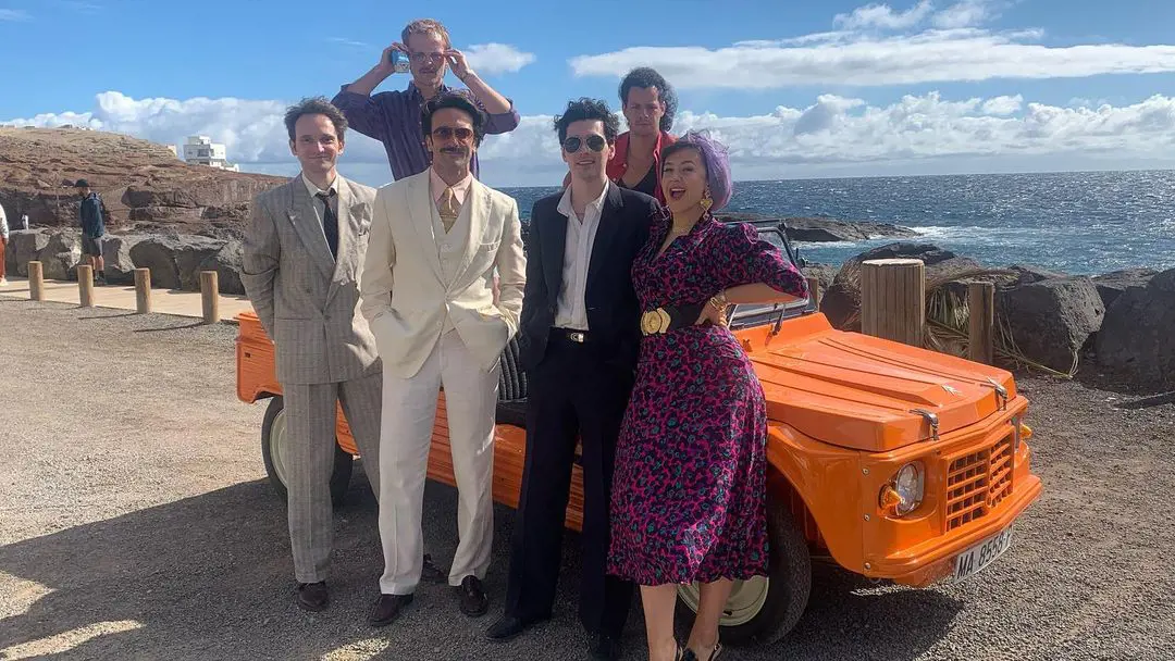 Some of the cast members of A Town Called Malice poses besides an orange car in a stunning backdrop